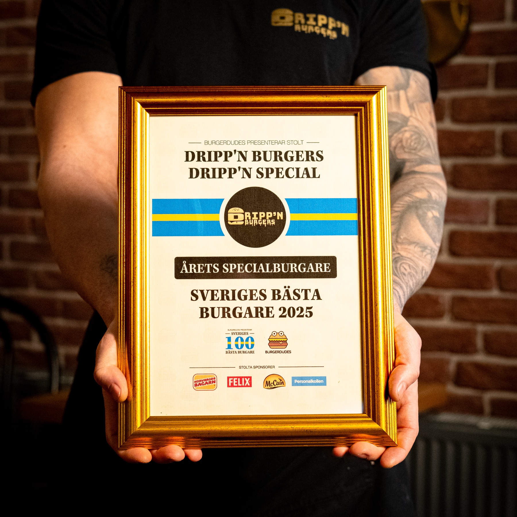 Diplomet från Burgerdudes hålls upp framför tegelväggen: Dripp'n Special, Årets Specialburgare, Sveriges 100 bästa burgare 2025